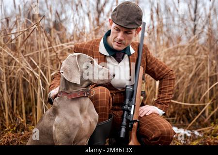 chasseur avec fusil de chasse en costume dans la forêt d'automne à la recherche de la proie trophée. beau homme caucasien avec arme et chiens de chasse traquer le g Banque D'Images