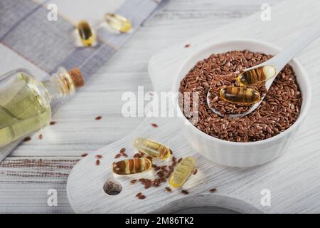 Brunir les graines de lin ou les graines de lin dans un petit bol et les capsules de gélatine avec de l'huile d'oméga sur fond de bois blanc. Banque D'Images