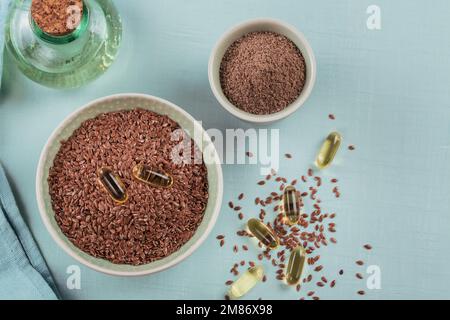 Brunir les graines de lin ou de lin et les graines de lin broyées ou broyées dans un petit bol et des capsules de gélatine avec de l'huile d'oméga sur fond bleu clair. Banque D'Images