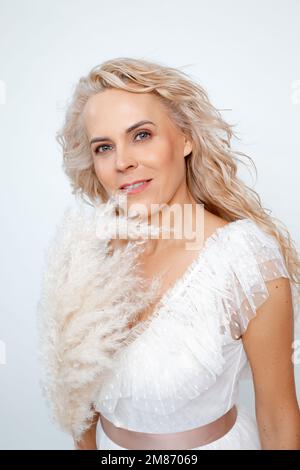 Souriante 50s femme adulte d'âge moyen avec une coiffure curly et des plumes décoratives en robe blanche isolée sur blanc, espace de copie. Ménopause légère Banque D'Images