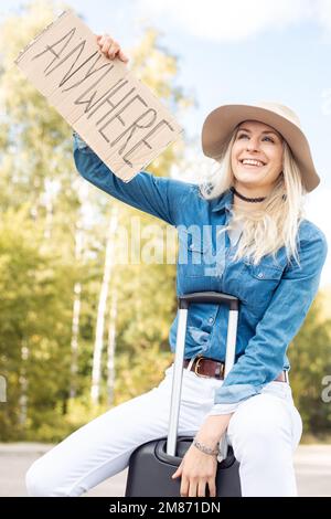 Une femme heureuse arrête de passer la voiture assise sur une valise avec une affiche en carton surélevé dans la forêt sur le bord de la route, de la vue du dessous. Lady in Hat échapper de la ville à Banque D'Images