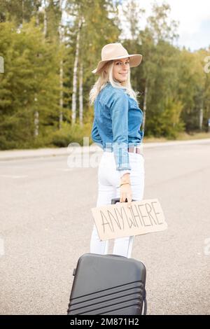 Une femme heureuse marche sur une route vide avec une affiche en carton et une valise, vue arrière. Une dame en chapeau et en denim s'évader de la ville en faisant un arrêt automatique pour aller Banque D'Images