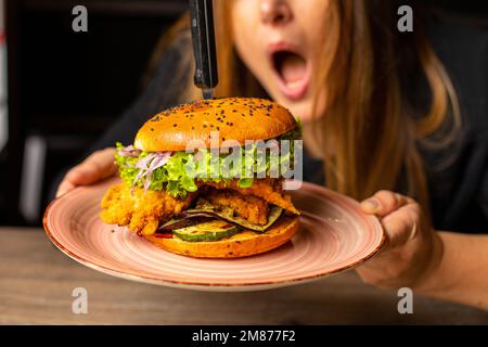 Photo de la femme choquée à bouche ouverte contenant un savoureux hamburger avec de la laitue, de l'oignon rouge, de la tomate et du poulet frit. Banque D'Images