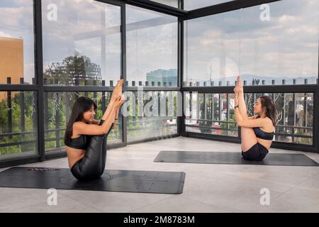 Deux belles jeunes femmes latines faisant des poses de yoga avancées dans le studio, portant des vêtements de sport, avec des fenêtres en arrière-plan. poses de yoga avancées. Banque D'Images