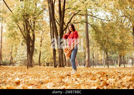 Femme souriante et élégante aux cheveux foncés, dans un foulard à carreaux chaud et un chandail rouge confortable qui pose et marche dans la forêt de feuilles dorées Banque D'Images