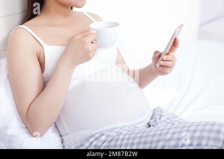 Une femme enceinte assise sur le lit a une boisson pendant l'utilisation des téléphones portables Banque D'Images