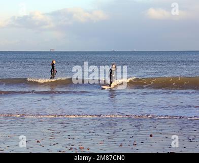 Deux jeunes femmes en combinaisons mouillées surfant sur de petites vagues près de la rive à Paignton, prises le 2023 janvier. Hiver Banque D'Images