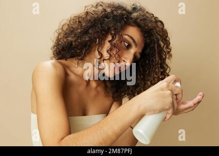 Femme applique de la crème et du baume à ses cheveux bouclés, le concept de protection et de soin avec des produits de salon, un look sain, un sourire aux dents sur un beige Banque D'Images