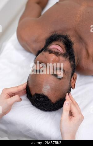 Spa thérapeute faisant un massage relaxant pour homme noir d'âge moyen. Un homme à barbe paisible se fait masser la tête dans un spa luxueux et moderne. Main Banque D'Images