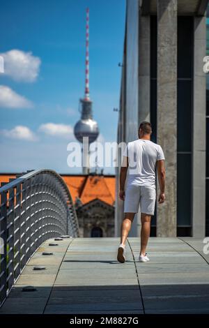 Un jeune homme blanc traverse Marie-Elisabeth-Lüders-Steg, un pont public piétonnier, avec la célèbre tour de télévision Alexanderplatz au loin Banque D'Images