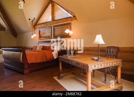 Bureau en bois antique avec chaise à haut dossier en cuir marron et lit traîneau dans la chambre principale à l'étage à l'intérieur de la luxueuse maison scandinave en rondins. Banque D'Images
