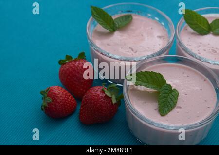 Gros plan de trois petits verres avec yaourt à la fraise à côté de quelques fraises et de feuilles de menthe sur une table bleue Banque D'Images
