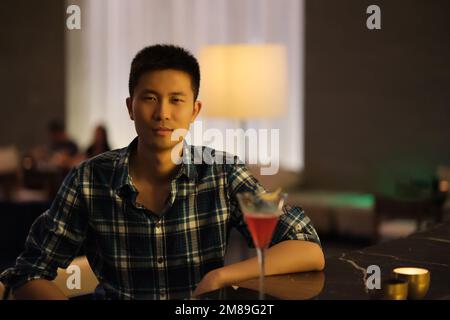 Portrait d'un beau jeune asiatique assis au comptoir du bar de nuit, avec un verre de cocktail, regardant l'appareil photo Banque D'Images