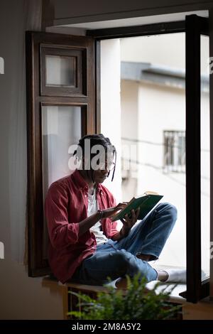 Un homme noir contrarié, excité par la lecture, est assis sur le rebord de la fenêtre avec des soucis de livre spectaculaires sur les personnages. Banque D'Images