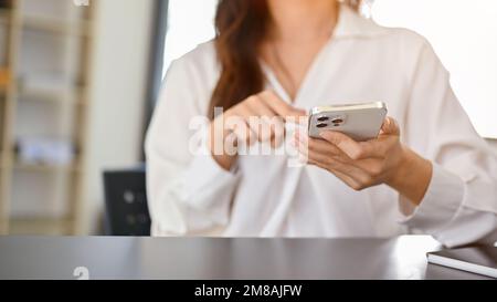 Jeune femme asiatique attirante vêtue de vêtements décontractés avec son smartphone, défilant sur les réseaux sociaux, recherchant quelque chose sur le site Web, discutant avec quelqu'un Banque D'Images