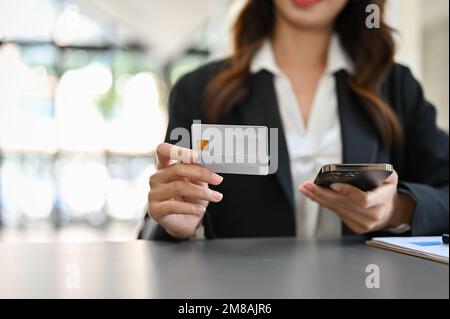 Image rognée, belle femme d'affaires asiatique se trouve à son bureau, tenant son téléphone et montrant sa carte de crédit à l'appareil photo. Banque D'Images