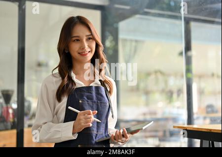 Une serveuse ou un café asiatique, jeune et sympathique, se tient debout dans son café, sous un tablier muni d'une tablette numérique. Banque D'Images