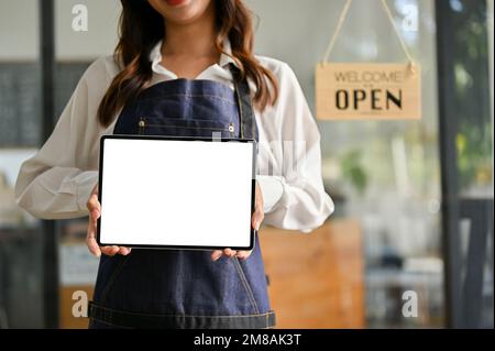 Belle jeune femme asiatique Barista ou serveuse en tablier se tient à la porte d'entrée de son café, tenant une tablette numérique, montrant une tablette blanche Banque D'Images