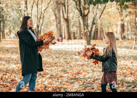 Vue latérale de la famille aimante de la jeune femme mère et fille adolescente tenant pile avec des feuilles dans la forêt en automne. Banque D'Images
