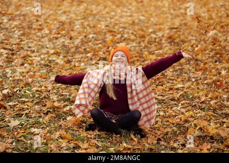 Bonne adolescente fille portant un chandail vineux, un chapeau orange, une écharpe à carreaux, assise sur des feuilles d'érable jaune. Banque D'Images