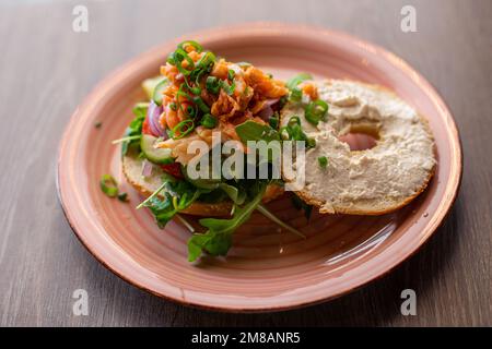 Vue latérale d'une assiette rose avec hamburger à dents ouvertes avec trou dans un petit pain, saumon, arugula, verdure, oignon vert, concombres. Banque D'Images