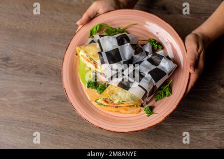 Photo rognée d'une femme tenant une grande assiette avec des burritos enveloppés dans du papier à carreaux avec des légumes décorés de légumes verts. Banque D'Images