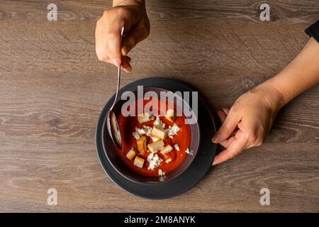 Vue de dessus d'une femme méconnaissable qui tient et fait fondre une soupe de tomates salées avec de la crème aigre et des croûtons dans un bol noir. Banque D'Images