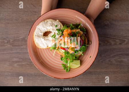 Photo rognée d'une femme tenant une assiette rose avec un hamburger plein de saveur avec trou dans un petit pain, saumon coupé, arugula, verdure. Banque D'Images