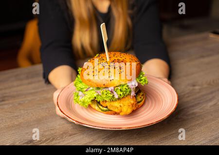 Photo rognée d'une femme assise à la table, avec assiette de hamburger frais avec laitue, oignon, tomate, poulet. Cuisson. Banque D'Images