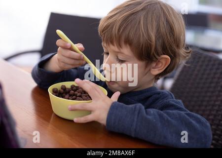 Petit garçon affamé assis à une table en bois dans la cuisine et prenant le petit déjeuner juste à côté. Enfant de l'âge de la maternelle tenant une cuillère pour manger des boules de chocolat sec Banque D'Images