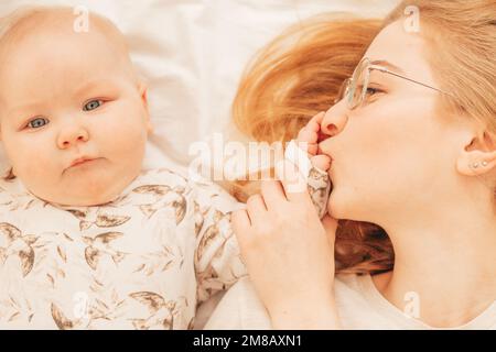 Une jeune mère gaie embrasse les doigts sur la paume du petit bébé allongé sur le lit de près. Photo de la famille à la maison de la femme et de l'enfant bébé mignon se reposant ensemble Banque D'Images