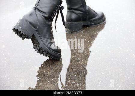 Jambes de femme en cuir noir bottes avec reflet dans la flaque dans une rue. Pluie avec neige en ville, hiver ou printemps, chaussures étanches Banque D'Images