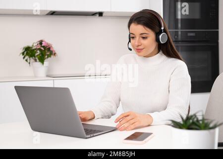 Le leader féminin du micro-casque discute des détails du projet avec l'équipe via Notebook. Jeune femme brune assis à table dans la cuisine travaillant en ligne au ralenti Banque D'Images