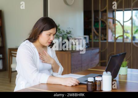 Femme enceinte asiatique consultant médecin en ligne sur ordinateur portable au sujet de son mal de gorge dans le salon de la maison Banque D'Images