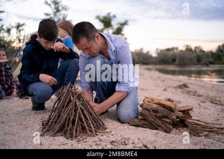 père et fils faisant un feu de camp le soir sur les rives du lac, papa enseignant à son fils adolescent comment faire un feu de camp. Copier l'espace Banque D'Images