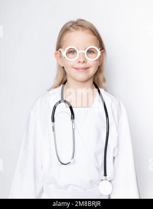 Portrait d'une petite fille mignonne vêtue d'une tenue de docteur avec stéthoscope et lunettes de jouet regardant l'appareil photo sur fond neutre Banque D'Images