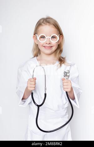 Portrait d'une petite fille mignonne vêtue d'une tenue de médecin avec des lunettes de jouet tenant le stéthoscope, souriant et regardant l'appareil photo sur un arrière-plan neutre Banque D'Images