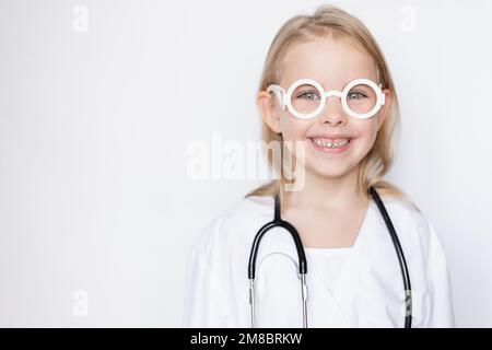 Portrait en gros plan d'une petite fille mignonne vêtue d'une tenue de médecin avec des lunettes de jouet tenant le stéthoscope, souriant et regardant l'appareil photo sur un dos neutre Banque D'Images