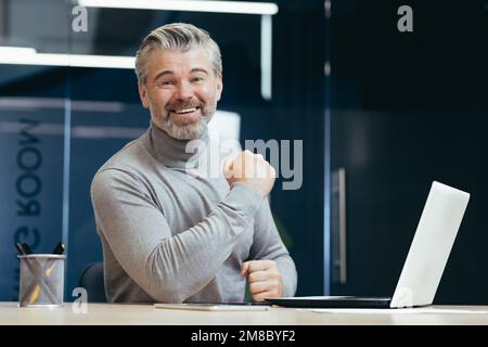 Portrait d'un homme d'affaires à la réussite aux cheveux gris. Un homme heureux est assis dans le bureau à la table avec un ordinateur portable. Célèbre la victoire, le succès, sourit à la caméra, montre Bravo. Banque D'Images