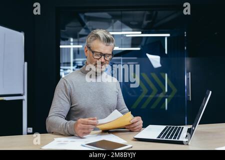 Homme senior aux cheveux gris travaillant au bureau avec un ordinateur portable et des documents. Il tient une enveloppe avec une lettre, des projets de loi et des rapports. Banque D'Images