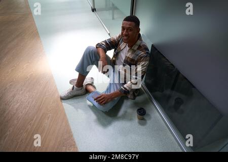 Homme afro-américain assis sur le sol, tenant le smartphone dans le bureau Banque D'Images