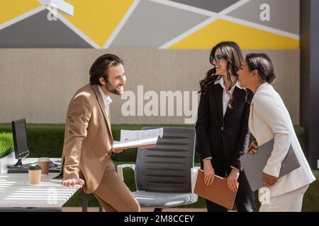 homme d'affaires souriant tenant des documents et parlant à des collègues multiethniques avec ordinateur portable et dossier dans le bureau, image de stock Banque D'Images