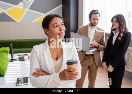 femme d'affaires multiraciale positive et réfléchie debout avec une tasse de papier près de collègues avec ordinateur portable sur un arrière-plan flou, image de stock Banque D'Images