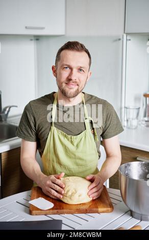 Caucasien gai mâle baker portrait dans le tablier vert uniforme sourire et regardant l'appareil photo avec la pâte sur le bois. Beau boulanger barbu à la maison assis à la table posant Banque D'Images