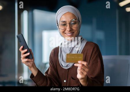 Femme d'affaires prospère dans le hijab travaillant à l'intérieur du bureau avec un ordinateur portable sur le lieu de travail, femme musulmane tenant le téléphone et la carte de crédit bancaire pour les achats en ligne et le transfert d'argent. Banque D'Images