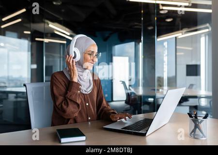 Femme d'affaires moderne prospère travaillant au bureau avec un ordinateur portable, femme musulmane portant le hijab et écouteurs écoutant des livres audio et des podcasts sur le lieu de travail, femme satisfaite et réussie. Banque D'Images