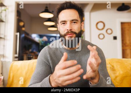 Portrait d'un jeune homme souriant à la barbe biraciale, en train de gesturer tout en étant assis sur un canapé dans la salle de séjour Banque D'Images