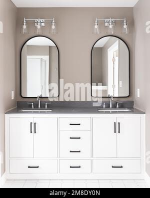 Salle de bains avec meuble-lavabo blanc et quincaillerie noire, robinets chromés sur comptoir en quartz, miroirs voûtés et murs bruns. Banque D'Images