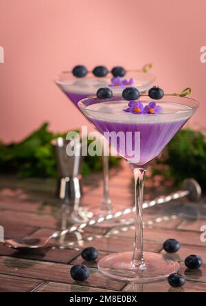 Un cocktail de gin violet avec un dessus en mousse garni de bleuets et de fleurs dans une scène de bar. Banque D'Images