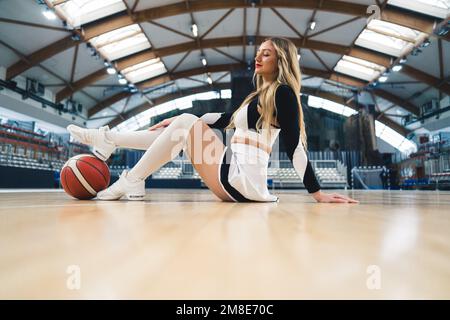 Un meneur s'assoit en toute confiance sur le sol avec son pied reposant sur le basket-ball. Vêtu d'une mini-jupe ludique associée à des chaussettes hauteur genou. Un terrain de basket-ball en arrière-plan peut être vu. Banque D'Images